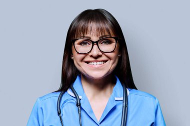 Headshot portrait of smiling middle-aged woman nurse in blue with stethoscope on grey studio background. Medical services, nhs, health, professional assistance, medical care concept clipart