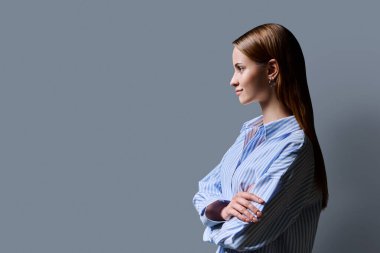 Kızıl saçlı güzel bir kızın profil portresi. Gri stüdyo arka planı. Kendine güvenen genç bir üniversite öğrencisi kollarını kavuşturarak boş alan arıyor. Eğitim gençliği