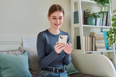 Akıllı telefon kullanan mutlu bayan ergen, evdeki koltukta oturan, 16,17-18 yaşındaki kız mesajları okuyor. İletişim, eğlence, öğrenme ve alışveriş için modern dijital teknolojiler