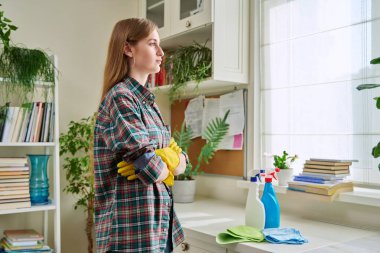 Portrait of young woman wearing protective rubber gloves for house cleaning, with arms crossed looking at window, in home room interior. Cleanliness, routine, lifestyle, youth concept clipart