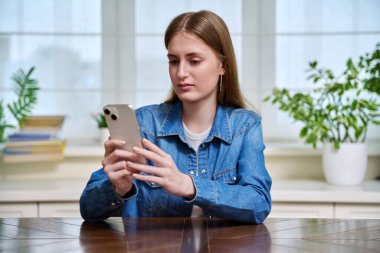 Akıllı telefon kullanan, evde oturan, 16-17-18 yaşlarında mesaj okuyan ciddi bir genç kız. İletişim, eğlence, öğrenme ve alışveriş için modern dijital teknolojiler