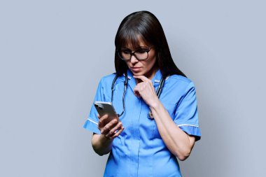 Ciddi bir bayan hemşire gri stüdyo arka planında akıllı telefon kullanıyor. Mobil uygulamalar, tıbbi hizmetlerdeki teknolojiler, sağlık, profesyonel yardım, tıbbi bakım kavramı