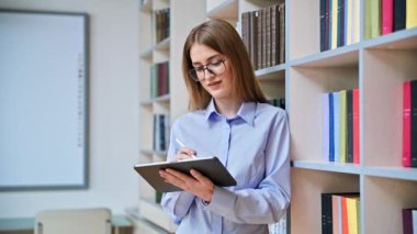 17-18 yaşlarında ciddi bir genç kızın portresi. Sınıfın kütüphanesinde dijital tablet kullanıyor. Gözlüklü sarışın genç kadın, eğitim kitaplarıyla dolu raflar.