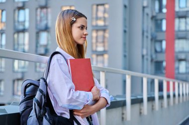 Sırt çantası kitaplarıyla gülümseyen üniversiteli genç bir kızın portresi açık havada kızıl saçlı, modern şehir geçmişi olan çekici bir genç kız. Gençlik, yaşam tarzı, eğitim kavramı