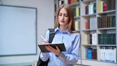 17-18 yaşlarında ciddi bir genç kızın portresi. Sınıfın kütüphanesinde dijital tablet kullanıyor. Gözlüklü sarışın genç kadın, eğitim kitaplarıyla dolu raflar.