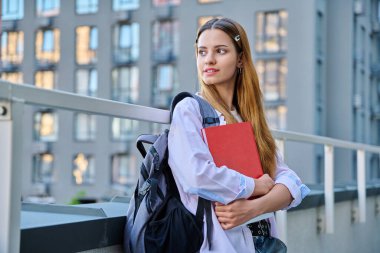 Sırt çantası kitaplarıyla gülümseyen üniversiteli genç bir kızın portresi açık havada kızıl saçlı, modern şehir geçmişi olan çekici bir genç kız. Gençlik, yaşam tarzı, eğitim kavramı