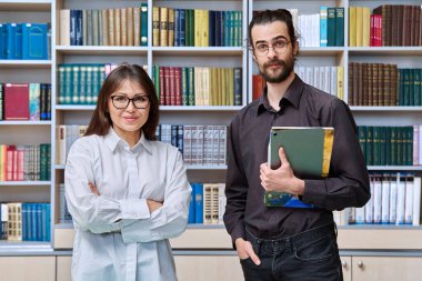 Erkek ve kadın meslektaşlarının portresi, eğitmenler, akıl hocaları, kitap rafları ve kütüphaneyle birlikte sınıfta birlikte okul danışmanlarına koçluk yapıyor. Kamera, takım çalışması ve takım konseptine bakan iki öğretmen.