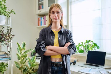 16-18 yaşlarında genç bir kızın portresi. Evde çapraz kollarla kameraya bakarak kendinden emin gülümsüyor. Uzun düz saçlı güzel sarışın kız. Gençlik yaşam tarzı güzellik modası