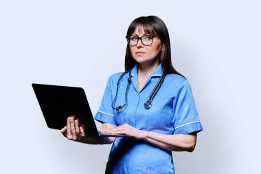 Ciddi bir bayan hemşire dizüstü bilgisayar tutuyor, beyaz stüdyo arka planındaki kameraya bakıyor. Mobil uygulamalar tıbbi hizmetler, sağlık, profesyonel yardım, tıbbi bakım konsepti teknolojileri