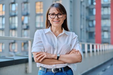 Çapraz kolları olan, açık havada poz veren, modern şehir gökdelenleri tarzı şehirli olgun, kendine güvenen bir kadının portresi. Yaşam tarzı, orta yaş, iş hayatı, başarı mutluluk kavramı