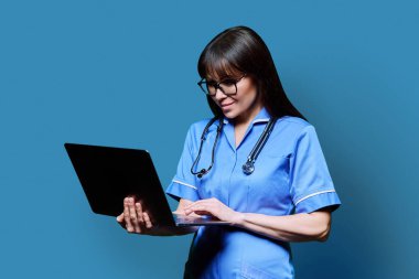 Gülümseyen kadın hemşire dizüstü bilgisayar kullanıyor, profil görüntüsü mavi stüdyo arka planında. Dijital uygulamalar, tıbbi hizmetler teknolojileri, sağlık, profesyonel yardım, tıbbi bakım konsepti