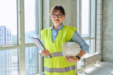 Sarı güvenlik yeleği giymiş, kasketli, kendine güveni tam bir kadın inşaat alanında kameraya bakıyor. Başarılı gülümseyen orta yaşlı kadın mühendis müfettişi, endüstri işi konsepti