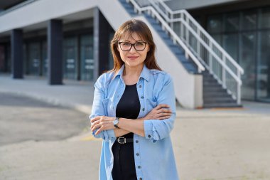 Açık havada gülümseyen, kendine güvenen, kollarını kavuşturup şehirde kameraya bakan olgun bir kadının portresi. Yaşam tarzı, orta yaş, iş, iş, insan kavramı