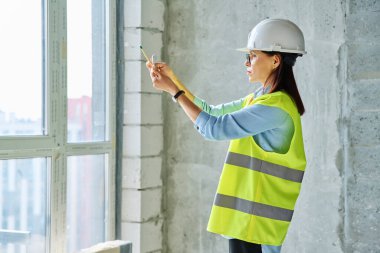 Profesyonel sanayi işçisi, inşaat alanında akıllı telefon kullanan koruyucu yelek giyen kadın mühendis tasarımcı denetçi. Endüstri, iş, insanlar, teknoloji kavramı