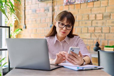 Orta yaşlı gülümseyen bir iş kadınının portresi dizüstü bilgisayarla çalışıyor, telefon kullanıyor, ofis içinde masa başında oturuyor. İş, uzaktan kumanda, serbest çalışma, teknoloji, 40 'lı yaşlarda insanlar