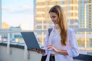 Genç, çekici bir üniversite öğrencisi dışarıda dizüstü bilgisayar kullanıyor, modern şehir geçmişi var. Eğitim, teknoloji, eğitim, 17-18 yaş gençlik kavramı