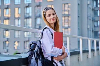 Sırt çantası kitaplarıyla gülümseyen üniversiteli genç bir kızın portresi açık havada kızıl saçlı, modern şehir geçmişi olan çekici bir genç kız. Gençlik, yaşam tarzı, eğitim kavramı