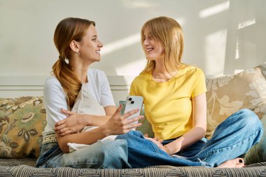 İki neşeli, gülen genç kız arkadaş evdeki kanepede akıllı telefonlarıyla oturup eğleniyorlar. Gençler gençlik eğlencesi yaşam tarzı arkadaşlık kavramı