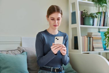 Akıllı telefon kullanan mutlu bayan ergen, evdeki koltukta oturan, 16,17-18 yaşındaki kız mesajları okuyor. İletişim, eğlence, öğrenme ve alışveriş için modern dijital teknolojiler