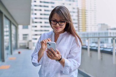 Akıllı telefon kullanan olgun, kendine güvenen bir kadın, modern şehir geçmişi. İş için mobil uygulama uygulamaları kullanan orta yaşlı başarılı bir kadın. Teknoloji internet işi, şehir tarzı
