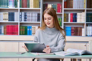 Genç kız öğrenci dijital tablet kullanarak eğitim kütüphanesinde okuyor. Eğitim, ergenlik, lise, üniversite, bilgi, öğrenim, teknoloji kavramı
