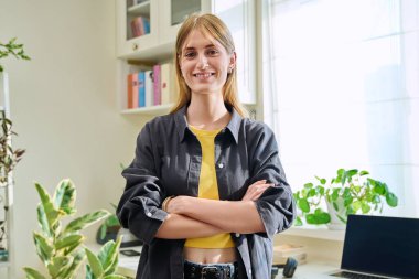 16-18 yaşlarında genç bir kızın portresi. Evde çapraz kollarla kameraya bakarak kendinden emin gülümsüyor. Uzun düz saçlı güzel sarışın kız. Gençlik yaşam tarzı güzellik moda konsepti