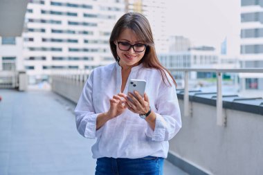 Akıllı telefon kullanan olgun, kendine güvenen bir kadın, modern şehir geçmişi. İş için mobil uygulama uygulamaları kullanan orta yaşlı başarılı bir kadın. Teknoloji internet işi, şehir tarzı