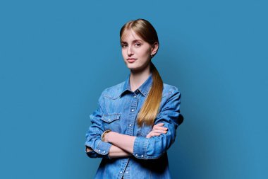 Sarışın, kendine güvenen genç kız kollarını kavuşturup mavi arka plandaki kameraya bakıyor. Başarılı genç kız öğrenci üniversite yaşı gençlik için reklam ürünleri ve hizmetleri