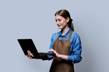 Önlüklü, laptopla gülümseyen, kendine güvenen genç bir kadının portresi. Gri stüdyo arka planındaki bilgisayar ekranına bakıyor. İşçi, girişim, küçük işletme, iş, hizmet sektörü, personel, gençlik kavramı