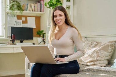 Dizüstü bilgisayarlı güzel bir kadın evdeki kanepede kameraya bakıyor. Gülümseyen çekici kadın bilgisayar kullanarak eğitim eğitimi eğlence iletişim teknolojisi gençlik yaşam tarzı eğitimi alıyor.