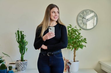 Duvarın yanında duran akıllı telefonu kullanan genç bir kadın. Kadın mesajlaşması okuma, mobil uygulama teknolojisi iş eğitimi için internet hizmetleri eğlence iletişim alışveriş yaşam tarzı