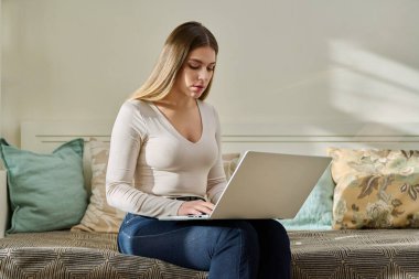 Evde çalışan genç bir kadın, dizüstü bilgisayar kullanarak kanepede oturuyor. Serbest meslek, eğitim, eğitim, uzaktan çalışma, boş zaman, internet teknolojisi uygulamaları konsepti