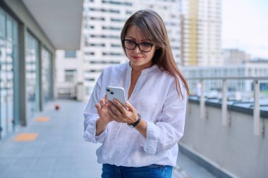 Akıllı telefon kullanan olgun, kendine güvenen bir kadın, modern şehir geçmişi. İş için mobil uygulama uygulamaları kullanan orta yaşlı başarılı bir kadın. Teknoloji internet işi, şehir tarzı