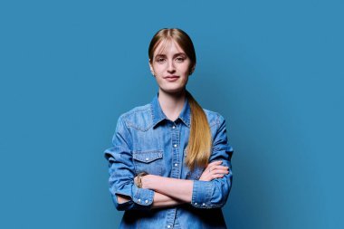Sarışın, kendine güvenen genç kız kollarını kavuşturup mavi arka plandaki kameraya bakıyor. Başarılı genç kız öğrenci üniversite yaşı gençlik için reklam ürünleri ve hizmetleri