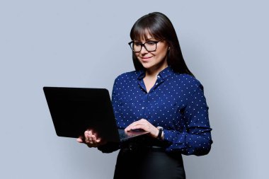 Orta yaşlı gülümseyen başarılı iş kadını portresi dizüstü bilgisayarla gri stüdyo arka planındaki bilgisayara bakıyor. İş, kariyer, teknoloji, başarı, insan kavramı.