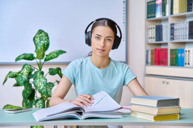 Genç kız öğrenci kulaklık takıyor lise kütüphanesinde sınav sınavlarına hazırlanıyor, kameraya bakıp gülümsüyor, ders kitapları ve kitaplarla oturuyor. Eğitim ergenlik bilgisi