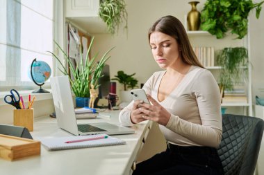Akıllı telefon kullanan, evinde oturan, 20-22 yaşlarında mesaj okuyan bir kadın. İletişim, eğlence, öğrenme ve alışveriş için modern dijital teknolojiler