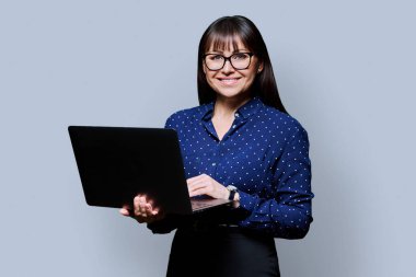 Orta yaşlı gülümseyen başarılı iş kadını portresi dizüstü bilgisayarla gri stüdyo arkaplanındaki kameraya bakıyor. İş, kariyer, teknoloji, başarı, insan kavramı.