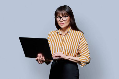 Orta yaşlı, kendine güvenen bir iş kadınının portresi dizüstü bilgisayarla gri stüdyo arkaplanındaki kameraya bakıyor. İş, kariyer, teknoloji, başarı, insan kavramı.