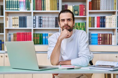 Kütüphanede çalışan genç erkek öğretmen akıl hocası portresi. Dizüstü bilgisayarla oturuyor. Yetişkin üniversite öğrencisi kameraya bakarak sınava hazırlanıyor. Eğitim öğretim teknolojisi