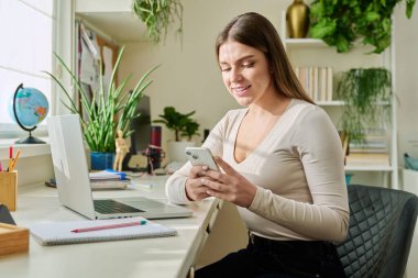 Akıllı telefon kullanan mutlu, gülümseyen genç bir kadın, evde oturan, 20-22 yaşlarında mesaj okuyan bir kadın. İletişim, eğlence, öğrenme ve alışveriş için modern dijital teknolojiler