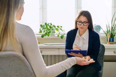 Orta yaşlı kadın psikoterapist genç bir kadınla seans terapisi ruh sağlığı uzmanı sosyal hizmet görevlisi olarak çalışıyor. Not panosuna not alıyor. Psikoterapi psikoloji danışmanlık desteği