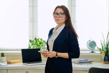 Orta yaşlı ciddi bir iş kadını elinde telefonla ofisteki kameraya bakıyor. Akıllı telefonlu, kendinden emin başarılı bir kadın. İş hayatı iletişimi için internet teknolojisi.