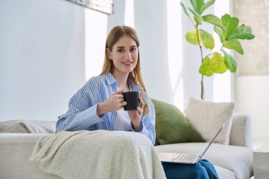 Dizüstü bilgisayarlı, bir fincan kahve içen, evdeki kanepede oturup kameraya bakan güzel, gülümseyen sarışın kadının portresi. Gençlik, yaşam tarzı konsepti