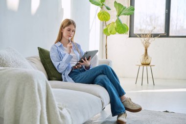 Evdeki kanepede dijital tablet kullanan genç bir kadın. Cihazlı kadın üniversite öğrencisi, rahatlama eğitimi, eğlence iletişimi eğitimi teknolojisi, yaşam tarzı gençlik konsepti