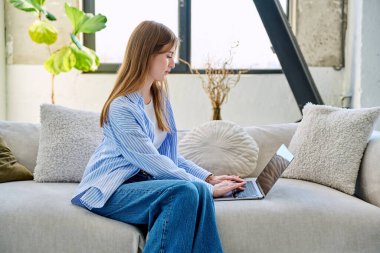 Genç bir kadının portresi kanepede oturuyor ve dizüstü bilgisayarda ev içinde daktilo yazıyor. Üniversite öğrencisi, internetten sosyal medya blogu alışverişini rahatlatmak için sınava hazırlanıyor.