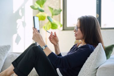 Evdeki kanepede oturup dijital tablet kullanan orta yaşlı güzel bir iş kadınının portresi, gevşemiş ciddi bir kadın. Teknoloji, yaşam tarzı, iş eğlencesi, insan konsepti