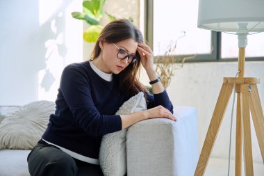 Olgun, üzgün, stresli bir kadın. Stresli, zihinsel ve fiziksel bir acı yaşıyor. Kanepede oturuyor. Sorunlar yalnızlık, baş ağrısı hormon stresi, orta yaşlı kadınların sağlığı