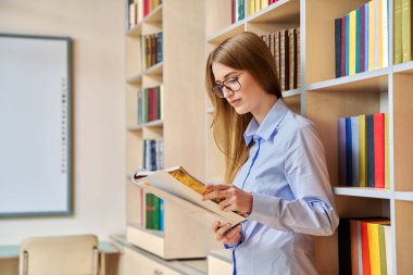 17-18 yaşlarında bir genç kızın portresi. Sınıf kütüphanesinde ders kitaplarını tutuyor. Eğitim kitapları arka plan görüşlü sarışın genç kadın rafları. Eğitim eğitimi öğrenimi