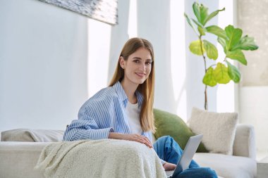 Dizüstü bilgisayarı olan güzel, gülümseyen sarışın bir kadının portresi. Evdeki kanepede oturmuş kameraya bakıyor. Gençlik, yaşam tarzı konsepti
